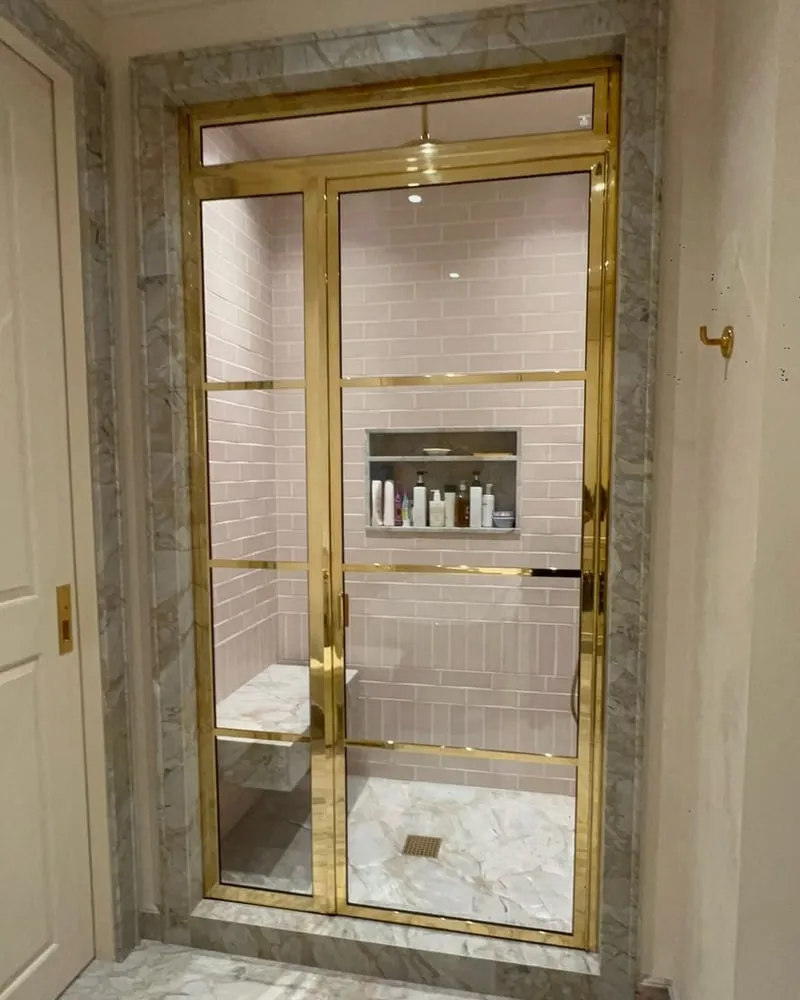 A brass grid-framed shower door on pink subway tile, framed in grey marble with a lit niche